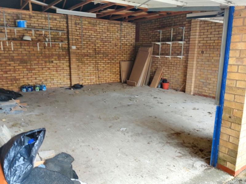 View into garage through the garage gate. On the floor are small debris and dust. In the bottom left on the foreground is a rubbish bag, and assorted trash, Against the wall in the back are old cans of paint. In the far corner are leftover plaster-boards. The wall has some shelves that are virtually empty. For a ceiling there are wooden beams, with one fluorescent tube light fixture across two of them. The walls are bare brickwork. On the rear wall, roughly 1.3 metres from the ground is a double power socket.
